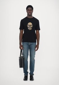 Zwarte t-shirt met schedelafbeelding, denim jeans, zwarte schoenen en een cilindrische zwarte tas met een hengsel. Model staat tegen een witte achtergrond.