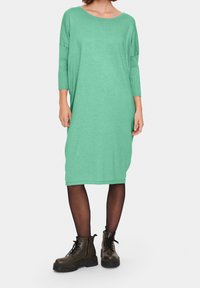 Grön, oversized stickad klänning med trekvartslånga ärmar och rundad halsringning. Buren med svarta tights och kraftiga bruna kängor.