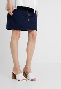 Woman wearing a navy button-front skirt and white woven high-heeled mules with wooden block heels on a plain background.