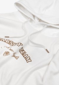 Hvid hættetrøje med snoretræk i halsen, prydet med et brunt trykt "Varsity Team" design og et dekorativt emblem i midten. Blød tekstur.
