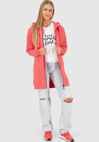 Korallfarbene Kapuzenjacke mit Reißverschluss und Taschen, kombiniert mit einem weißen T-Shirt mit schwarzem Text. Tragen mit hellblauen, zerrissenen Jeans und roten Turnschuhen.
