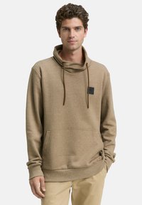 Bruine hoodie gemaakt van een zachte stof, met een hoge kraag, trekkoorden en een patch op de borst. Kangoeroezak en geribbelde manchetten.