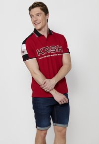 Piros poloing fekete és fehér kiegészítőkkel, a frontján merész "KRSH" és "UNLIMITED BRAND 2017" felirattal. Farmer rövidnadrággal viselve.