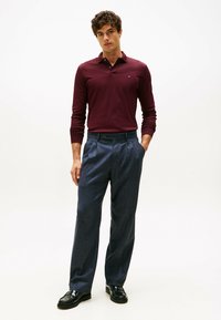 Langärmliges, burgunderfarbenes Poloshirt, kombiniert mit maßgeschneiderten, navyblauen Wollhosen mit glatter Textur und klassischer Passform. Schwarze Slipper runden den Look ab.
