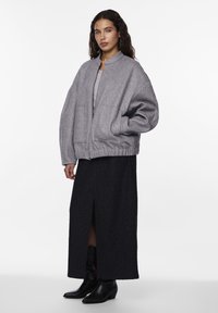 Kvinna i en grå oversized zip-up jacka, svart midikjol med framfåra, svarta klackstövlar och hoopörhängen mot en vit bakgrund.
