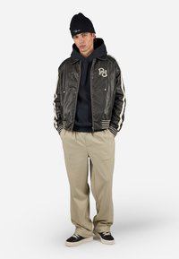 Zwarte leren bomberjack met witte strepen, geborduurd logo, gebreide marineblauwe sweatshirt, beige broek en zwart-witte schoenen.