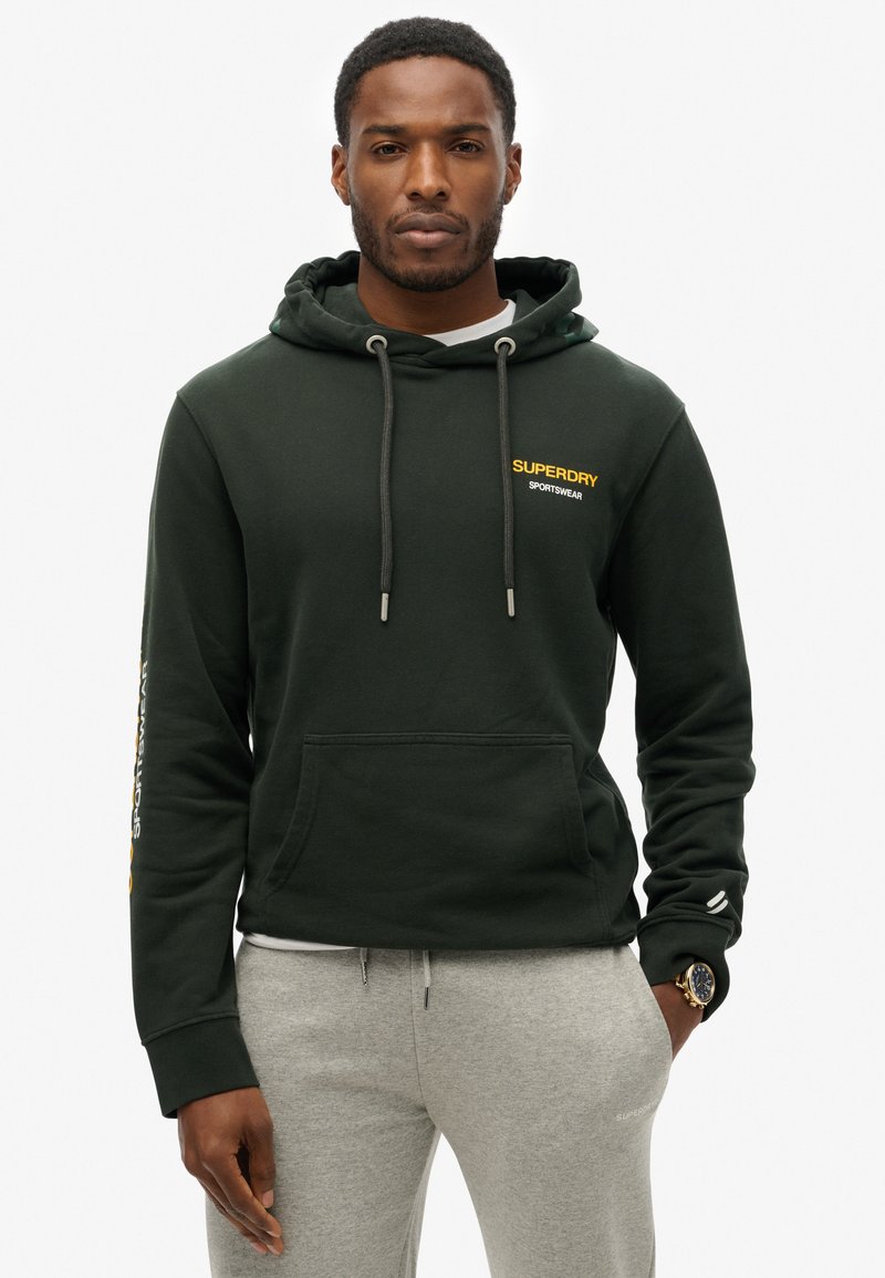 Felpa verde scuro realizzata in tessuto morbido, con tasca frontale, cappuccio con lacci e logo giallo sul petto e sul maniche. Indossata con pantaloni grigi.