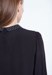 Blusa nera in tessuto strutturato, colletto decorato con perle, morbide pieghe sulle spalle e chiusura a bottone sul retro.
