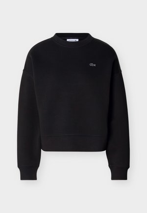 Crna majica s okruglim izrezom i dugim rukavima, s malim bijelim izvezenim Lacoste krokodilom na lijevoj strani prsnog koša.