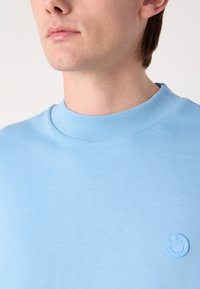 Světle modrý svetr s žebrovaným límcem. Obsahuje malé vyražené logo na hrudi. Měkká textura a ležérní design.