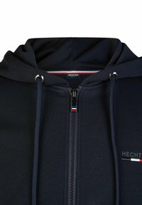 Marineblaue Zip-Up-Hoodie mit Kordelzügen, ausgestattet mit einem Metallreißverschluss und einem Logo auf der Brust. Angenehme Textur, lässiges Design und Kapuzenstil.