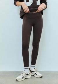 Noge v temno rjavih oprijetih pajkicah, bele nogavice s črnimi črtami ter bež in črni supergi z vezalkami na betonski podlagi.