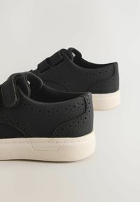 Schwarzer Sneaker mit perforierten Details, Klettverschluss und einer cremefarbenen Gummisohle; glatte Oberfläche mit einem abgerundeten Design.