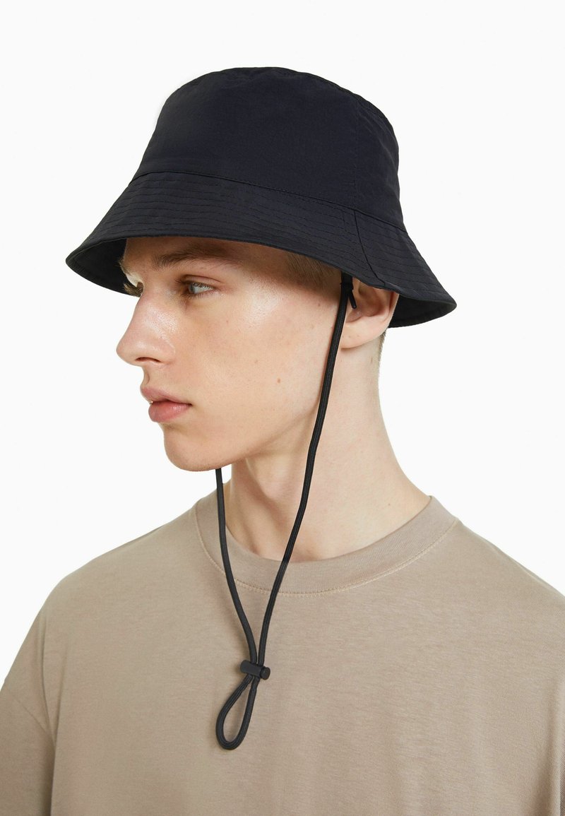 Sombrero negro de cubo con ala ancha y tela texturizada. Presenta una correa para la barbilla asegurada con un regulador. Se lleva con una camiseta beige de cuello redondo.