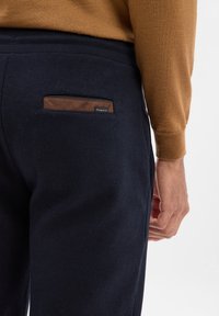 Granatowe dresy z teksturowanej tkaniny, z brązowym skórzanym akcentem w kieszeni i czarną metką. Prosty i funkcjonalny design.