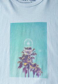 Vaaleansininen lyhythihainen t-paita, jossa on graafinen kuvasto palmupuista vihreän gradienttitaustan edessä, ja pyöreä valkoinen etiketti, jossa lukee "SUNSHADE."