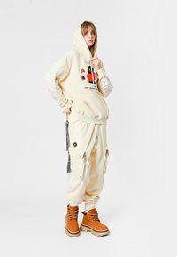 Mujer joven vistiendo pantalones de chándal color crema, sudadera a juego con un gráfico colorido y botas de encaje en tono marrón, de pie frente a un fondo blanco.