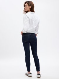 Sötétkék skinny farmernadrág, két hátsó zsebbel, fehér gombos inggel párosítva. A modellen fekete-fehér tornacipő van.