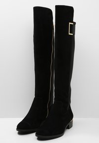 Black knee-high boots made of suede with a gold zipper detail, rounded toe, and a low block heel. Smooth texture with a sleek design.