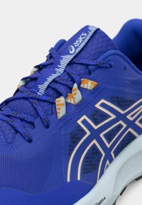 Μπλε αθλητικό παπούτσι με επάνω μέρος από διχτυωτό υλικό, αντίθετο πορτοκαλί λογότυπο και λεπτομέρειες, υφή επιφάνειας και επίπεδα κορδόνια. Ελαφρύ με μαλακή σόλα.