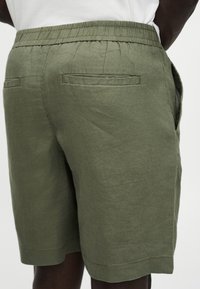Shorts à taille élastique vert olive avec poches passepoilées à l'arrière portés par une personne en chemise blanche, vus de la taille arrière jusqu'au milieu de la cuisse.