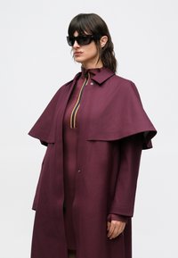Burgundy coat with a cape design, made of textured fabric. Features a stand collar, button closure, and a yellow striped accent at the neckline.