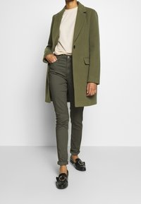 Abrigo largo verde oliva con solapas de muesca, camiseta de cuello redondo beige, pantalones ajustados verdes oliva y mocasines negros con detalle de herrajes circulares.