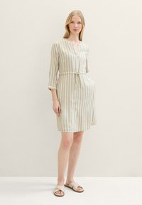 Robe chemise rayée en beige et crème, avec des manches 3/4 et une taille à nouer. Accompagnée de sandales beiges à lanières croisées.