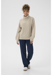 Beige kauluspusero, jossa on joustovyötärö, yhdistetty tummansiniset suorat housut ja vaaleat lenkkitossut yksinkertaisella taustalla.