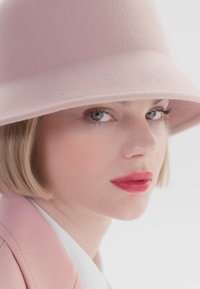 Rosa filt hatt med bred skjerm, myk tekstur og rund krone. Modell har en nøytral antrekk og lett sminke.