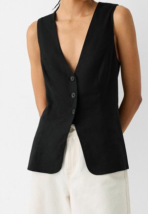 Person wearing a fitted sleeveless black vest with three buttons over light-colored pants against a plain background.