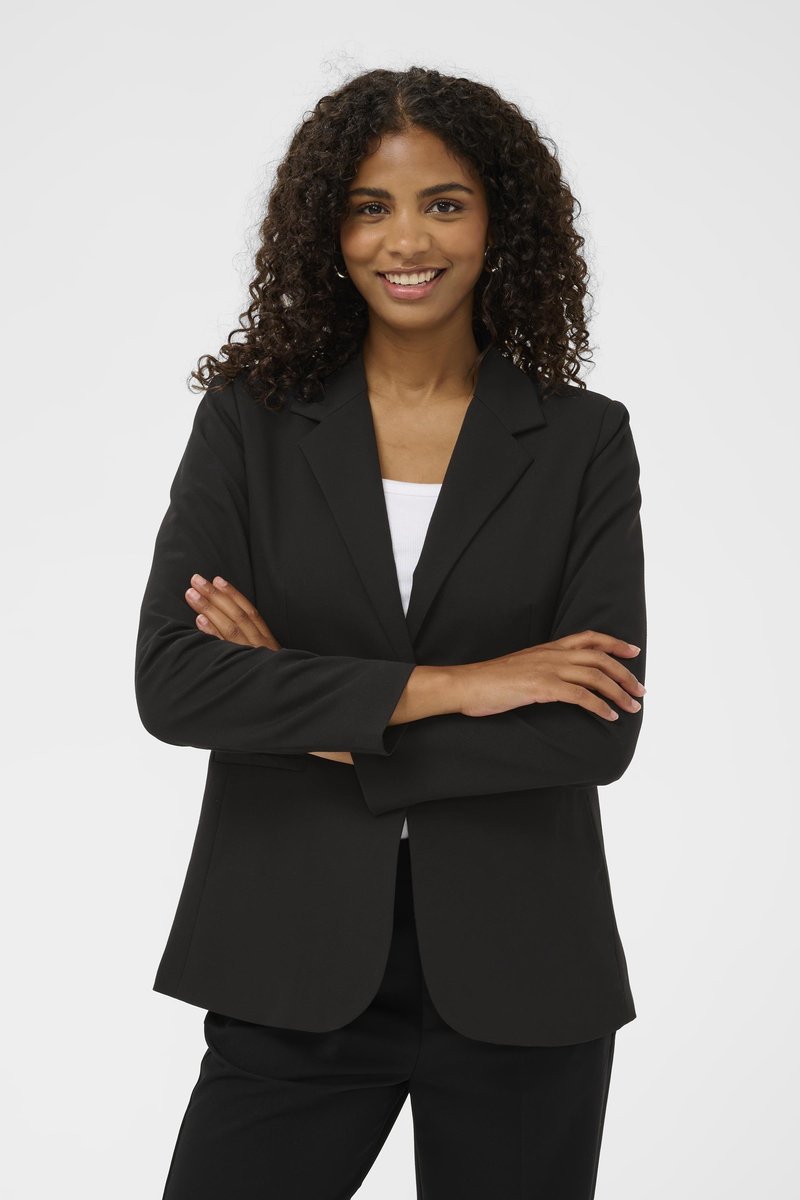 Usměvavá žena s kudrnatými vlasy, oblečená v černém saku a kalhotách, s bílým topem pod tím, stojící se založenýma rukama na jednobarevném pozadí.