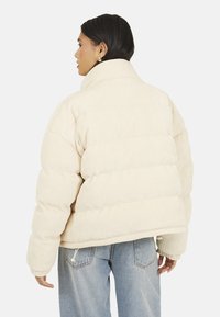 Pikowana ecru kurtka o prążkowanej fakturze, z wysokim kołnierzem i elastycznymi mankietami. Posiada regulowany sznurek u dołu i krótki fason.