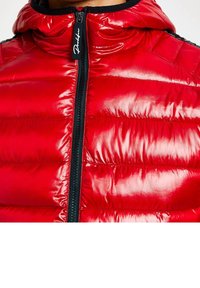 Veste rembourrée rouge avec une finition brillante, col haut et zip noir. Elle présente des coutures horizontales et une tirette de fermeture éclair estampillée.