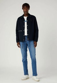 Tamsiai mėlynas švarkas su sagomis priekyje ir užtrauktuko kišene, dėvimas ant balto marškinėlio ir šviesiai mėlynų džinsų, derinamas su baltais sportbačiais.