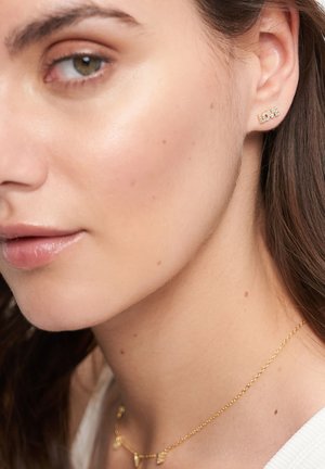 Kvindeansigt i profil iført en gylden ørestik med teksten "LOVE" og en delikat guldkæde med små bogstavsvedhæng.