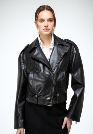 Jonge vrouw met een zwarte leren motorjas over een wit overhemd met kraag, staand tegen een eenvoudige lichte achtergrond.