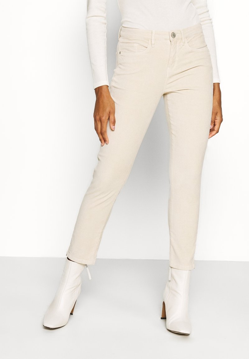 Femme portant un pantalon beige slim, un haut blanc à manches longues et des bottines blanches, debout devant un fond blanc.