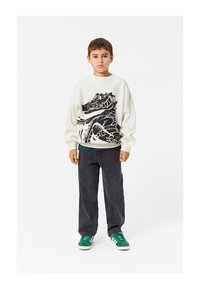 Felpa chiara con un design grafico nero, abbinata a pantaloni grigio scuro e sneakers verdi con strisce bianche.
