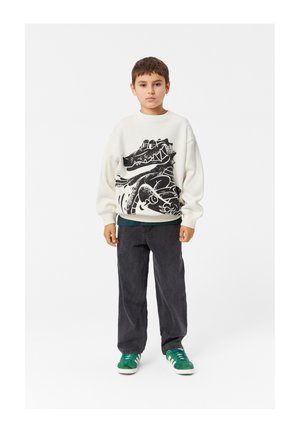Felpa chiara con un design grafico nero, abbinata a pantaloni grigio scuro e sneakers verdi con strisce bianche.