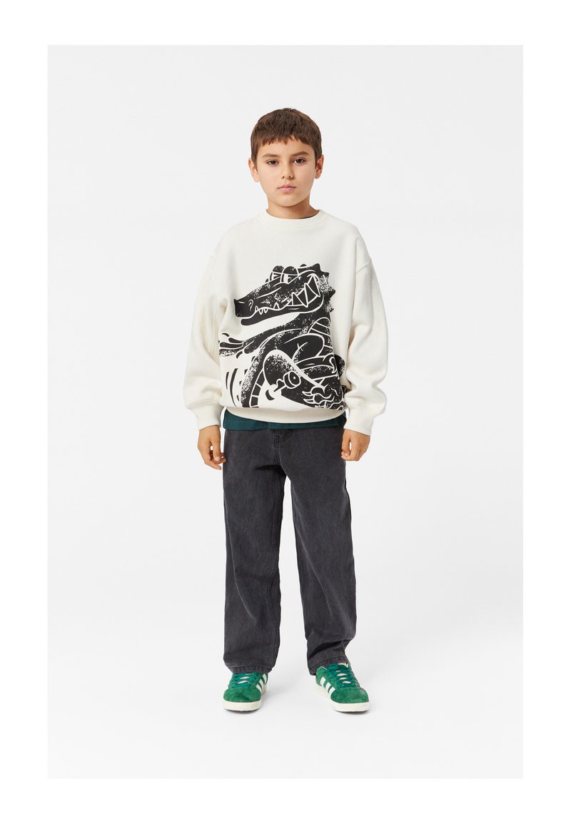 Felpa chiara con un design grafico nero, abbinata a pantaloni grigio scuro e sneakers verdi con strisce bianche.