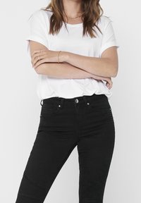 Donna che indossa una camicia bianca a maniche corte e pantaloni neri a vita alta, con le mani incrociate in vita; visibile un semplice bracciale e una collana d'oro.