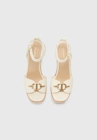 Sandales à talons hauts blanches avec une large bride et des détails en métal doré. Présente une fine bride de cheville et une semelle intérieure rembourrée.