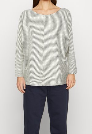 Woman wearing a white long-sleeve top with thin black chevron stripes and navy pants, standing against a plain light background.