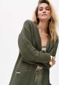 Olijfgroene gebreide cardigan met een gemêleerd patroon, open voorkant en twee voorzakken. Bevat een merklabel aan de zijkant.