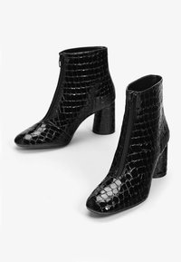 Botas negras hasta el tobillo con un patrón de cocodrilo, acabado brillante, punta afilada y cierre de cremallera en la parte delantera. Diseño de tacón alto grueso.