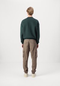 Man staat met zijn rug naar ons toe, gekleed in een donkergroen sweatshirt, bruine cargobroeken en lichtbeige sneakers, tegen een effen witte achtergrond.