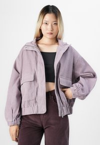 Jonge vrouw met steil blond haar, gekleed in een lila corduroy jas, een zwart crop top en donkere paarse corduroy broek tegen een witte achtergrond.