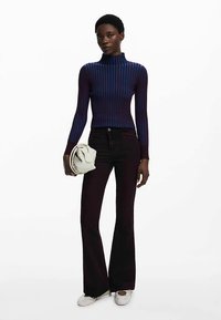 Pull à col roulé violet foncé à rayures bleues, associé à un pantalon évasé foncé et tenant un sac à main crème. Chaussures blanches.