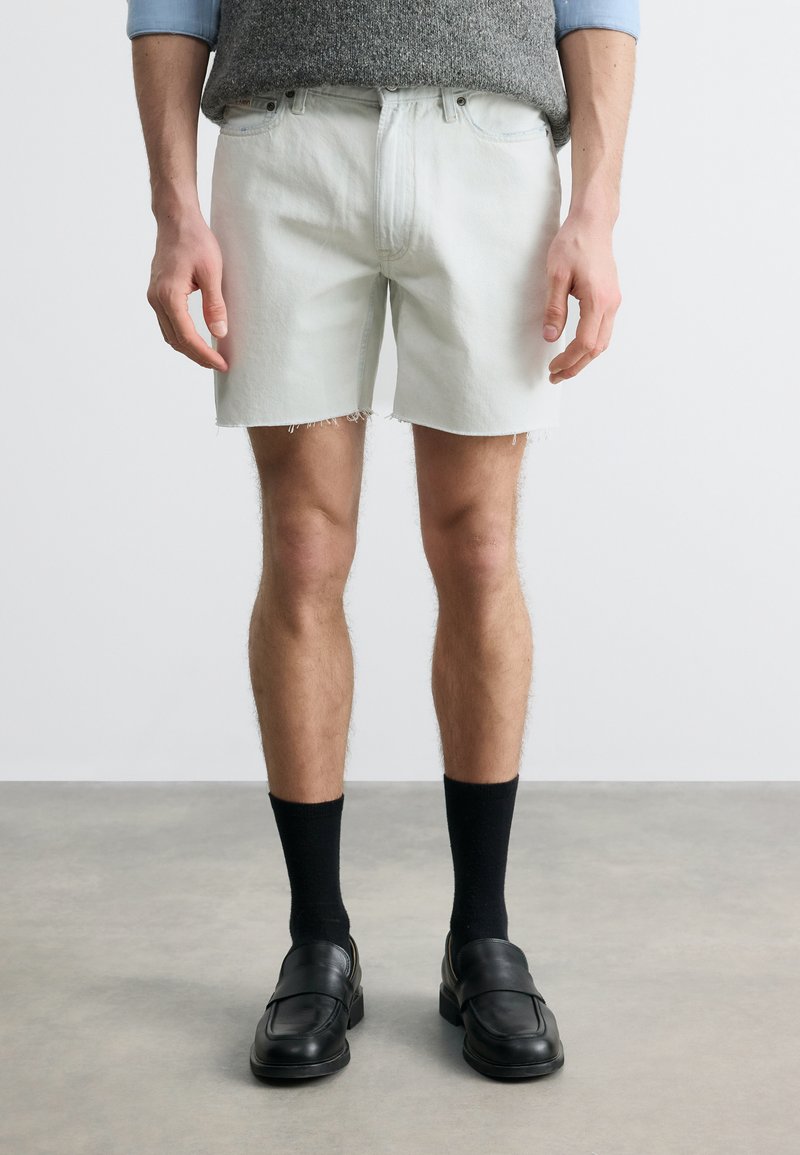 Άτομο που φοράει άσπρο τζιν σορτς με φθαρμένες άκρες, μαύρες κάλτσες, μαύρα δερμάτινα παπούτσια και γκρι πουλόβερ ορατό στη μέση.